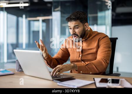Homme d'affaires regardant surpris et stressé tout en recevant de mauvaises nouvelles ou un problème inattendu sur son ordinateur portable, exprimant l'étonnement avec la bouche ouverte et répandre le geste de la main dans une configuration de bureau moderne Banque D'Images