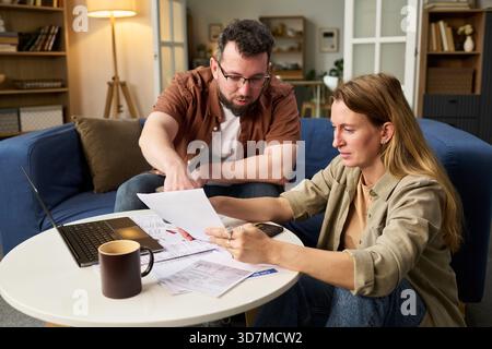 Homme d'âge moyen caucasien et femme d'âge moyen caucasien examinant les documents fiscaux ensemble à table, l'homme pointant le papier tandis que la femme tenant les documents, ordinateur portable et calculatrice visibles Banque D'Images
