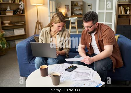 Femme jeune adulte caucasienne utilisant un ordinateur portable tandis que jeune homme adulte caucasien examinant des documents financiers à table, les deux collaborant sur le paiement des impôts avec la calculatrice et la paperasse Banque D'Images