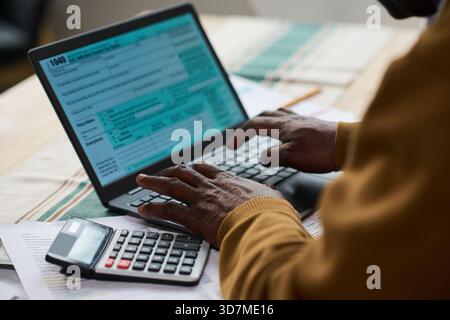Homme noir d'âge moyen à l'aide d'un ordinateur portable et d'une calculatrice tout en remplissant des formulaires d'impôt au bureau, les mains tapant sur le clavier avec des documents fiscaux et des documents financiers étalés sur la table Banque D'Images