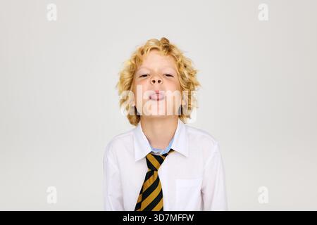 Un garçon blond joyeux dans une chemise blanche et une cravate rayée sort ludique de sa langue. Banque D'Images