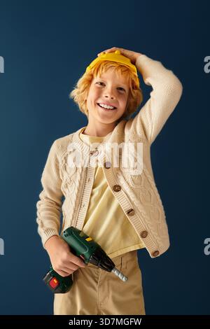Un garçon blond sourit en tenant un outil, portant des vêtements décontractés et un casque jaune dans un studio. Banque D'Images