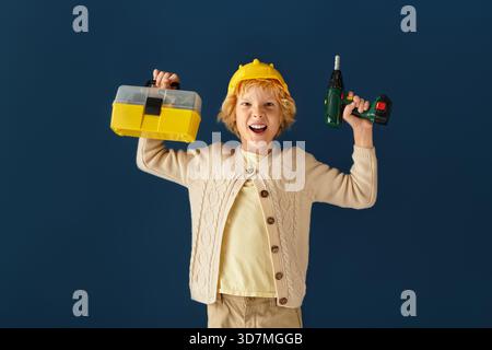 Un garçon blond joyeux avec les cheveux ondulés tient une trousse à outils et une perceuse dans un cadre de studio. Banque D'Images