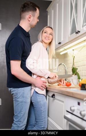 Couple cuisiner ensemble, profiter du temps dans la cuisine à la maison pendant l'escapade de week-end. Femme, amoureuse, hache des légumes dans la cuisine, tandis que l'homme l'embrasse de Be Banque D'Images