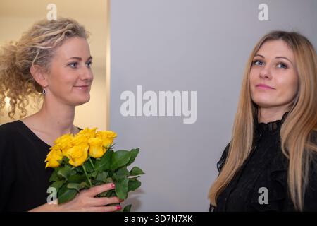 Deux femmes, dont une aux cheveux bouclés tenant des roses jaunes, partagent ensemble un moment joyeux, célébrant l’amitié et le bonheur dans un ensemble lumineux et accueillant Banque D'Images