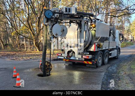 Un camion de nettoyage d'égout Hydro-vac enlève les débris d'un pipeline souterrain à travers un trou d'homme ouvert, avec des cônes de sécurité placés sur une route forestière tranquille Banque D'Images