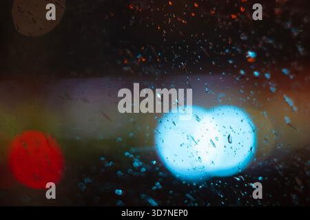 Une photo abstraite de lumières bokeh bleues et rouges vues à travers une surface de verre avec des gouttes de pluie. Banque D'Images