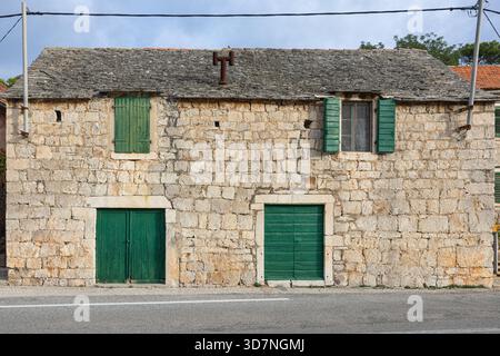 Une maison en pierre altérée avec quatre volets verts en bois se trouve dans une rue calme. La façade rugueuse, la petite porte et les supports à vélos en métal évoquent le charme rustique Banque D'Images
