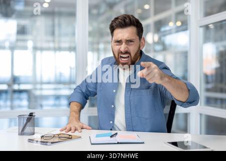 Homme en colère criant et pointant un doigt assis à un bureau dans un bureau moderne, exprimant la frustration, l'agressivité, le stress et l'insatisfaction dans un environnement de travail Banque D'Images