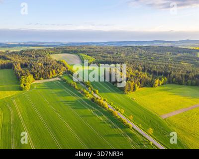 Des champs verdoyants et des forêts s'étendent à travers le paysage tandis que le soleil se couche un soir de printemps, projetant une lumière chaude sur le paysage vibrant. Les collines douces créent une toile de fond sereine. Banque D'Images