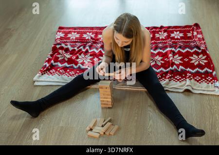 Fille empilant des blocs de bois sur une couverture confortable. Une adolescente est assise sur le sol avec une couverture festive, jouant un jeu d'empilement en bois avec focus et Banque D'Images