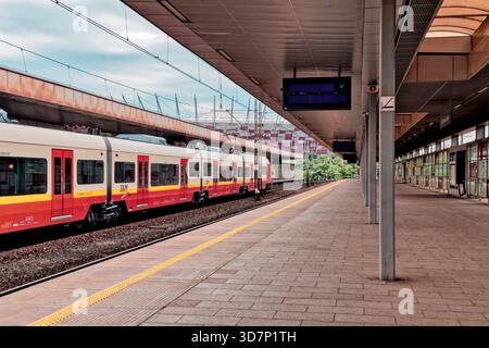 Le train SKM rouge et jaune attend à la gare de Warszawa Stadion, avec le stade national visible en arrière-plan par une journée calme et nuageuse. Banque D'Images