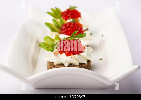 Trois délicieux canapés recouverts de crème, de caviar coloré et de légumes frais sont présentés sur une assiette blanche élégante. Cet élégant apéritif est Perfe Banque D'Images