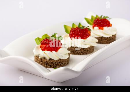 Trois délicieux canapés sont disposés sur un plat blanc avec une croûte de pain noir surmontée de crème, de caviar et d'herbes fraîches. Parfait pour n'importe quel festival Banque D'Images