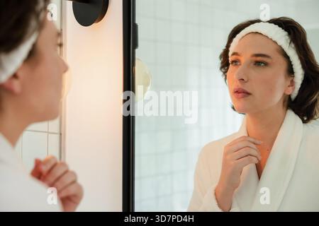 Examen de la peau dans un miroir à cadre noir et peignoir dans la salle de bains à la lumière du meuble-lavabo Banque D'Images