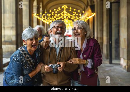 Groupe d'heureux amis seniors profitant de vacances ensemble, riant et s'amusant tout en explorant une arche de la ville décorée avec une lueur festive Banque D'Images