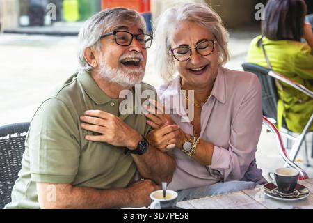 Couple mature riant et discutant autour d'un café sur un patio de café extérieur ensoleillé, appréciant la conversation détendue, la compagnie et la retraite insouciante Mo Banque D'Images