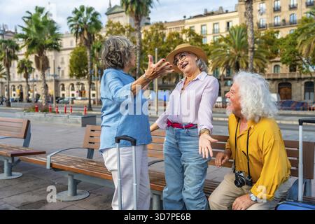 Groupe diversifié d'amis seniors heureux profitant de leurs vacances en ville ensemble, riant et communiquant sur un banc avec leurs bagages, représentant ACT Banque D'Images