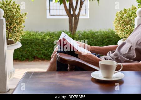 Une personne profite d'une matinée tranquille en lisant un livre tout en sirotant un café sur un patio. Une végétation luxuriante entoure la région, offrant une atmosphère sereine parfaite Banque D'Images