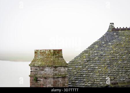 Cheminée en briques altérées et toit en ardoise couvert de mousse sur une matinée côtière brumeuse à Moint Saint Michel, en Normandie. Vieux toit en ardoise et cheminée vieillie. Banque D'Images