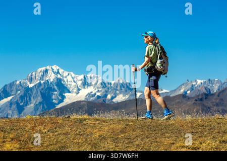 Un randonneur avec des bâtons de trekking marche sur une colline herbeuse avec des montagnes enneigées en arrière-plan. Val d'Aoste, Italie Banque D'Images