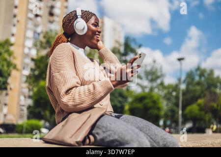 Portrait d'une femme noire joyeuse d'une vingtaine d'années utilisant son téléphone portable à l'extérieur dans un cadre urbain, portant des écouteurs sans fil et souriant pendant qu'elle écrit Banque D'Images