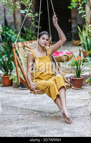 Femme en salopette jaune se détend sur une balançoire en bois dans un cadre de jardin luxuriant. Sierra Alhamilla, Almeria, Espagne Banque D'Images