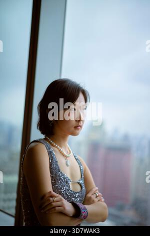 Jeune femme en haut pailleté regarde par une grande fenêtre sur un horizon urbain. Shanghai, Chine Banque D'Images