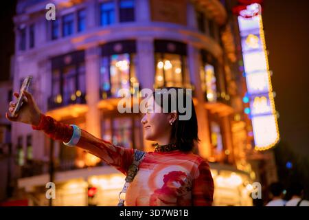 Femme prenant un selfie la nuit devant un bâtiment urbain très éclairé. Shanghai, Chine Banque D'Images