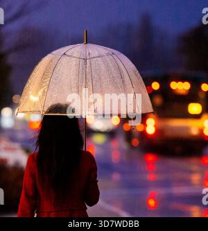 Femme en manteau rouge tenant un parapluie clair dans la rue pluvieuse et illuminée de la ville au crépuscule. WA, ÉTATS-UNIS Banque D'Images