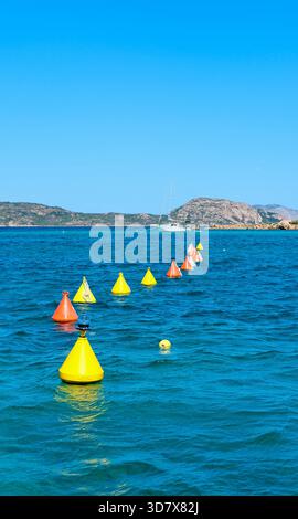 Série de bouées lumineuses sur la mer bleue ; côte rocheuse lointaine et voilier ancré visible ; tourné en plein jour avec un mouvement d'eau doux. Banque D'Images