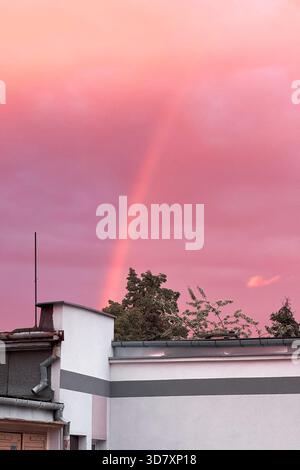 Plan vertical saisissant capturant un arc-en-ciel faible sur un ciel rose et violet spectaculaire, avec le sommet d'un bâtiment et des arbres luxuriants au premier plan. Banque D'Images