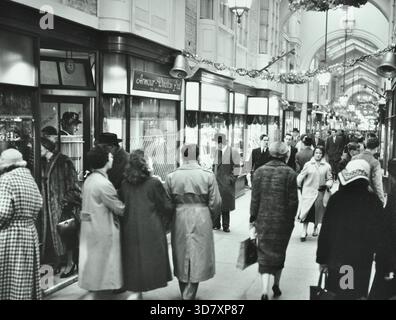 Burlington Arcade, Burlington Gardens, Westminster, Londres, 1960. Vue sur les boutiques orientées vers le sud depuis Burlington Gardens. Érigée en 1818-19 à partir des plans de Samuel Ware, la galerie marchande de luxe couverte de 179 mètres de long a été construite sur les anciens jardins de Burlington House, la maison de George Cavendish, le 1er comte de Burlington. Les panneaux vitrés du toit-lampadaire sont soutenus par des arches, qui divisent les 72 unités d'origine de deux étages en baies de deux magasins avec des fenêtres d'affichage en saillie. Une ligne de lanternes peut être vue suspendue au toit avec des décorations de Noël suspendues. À gauche de Banque D'Images