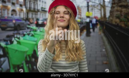 Femme utilise son doigt pour pointer la rue animée tandis qu'une jeune voyageuse blonde dans un béret rouge et des lunettes explore une scène urbaine. Banque D'Images