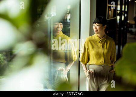 Une femme portant une blouse jaune éclatante se tient gracieusement près d'une grande fenêtre. Elle sourit doucement, reflétant un comportement confiant. Lumière naturelle améliorée Banque D'Images