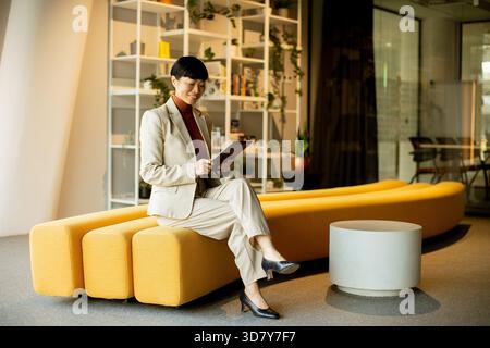 Un professionnel des affaires est assis sur un banc jaune dans un bureau contemporain. Elle tient une planche à pince, portant un costume léger et souriante, entourée d'étagères Banque D'Images