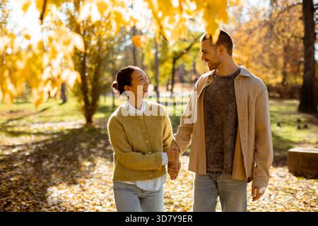 Un couple marche main dans la main à travers un parc rempli de feuilles d'automne colorées. Ils se sourient l'un à l'autre, se prélassant dans la lumière chaude du soleil. Banque D'Images
