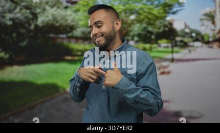 Homme, jeune dans les haussements de chemise en denim avec les paumes vers le haut, les mains visibles et souriant dans une rue par une passerelle verte de parc ; incertitude ludique. Banque D'Images