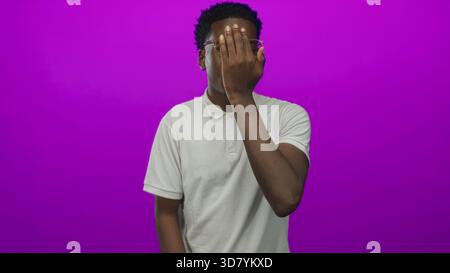 Homme couvrant l'oeil avec la main dans le studio violet portant des lunettes sous une lumière vive douce ; intrigue mystérieuse. Banque D'Images