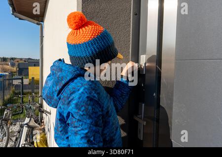 Garçon en veste d'hiver bleue et bonnet tricoté avec pompon déverrouillage porte d'entrée avec clé le jour ensoleillé. Concept d'indépendance de l'enfance, retour à la maison fr Banque D'Images