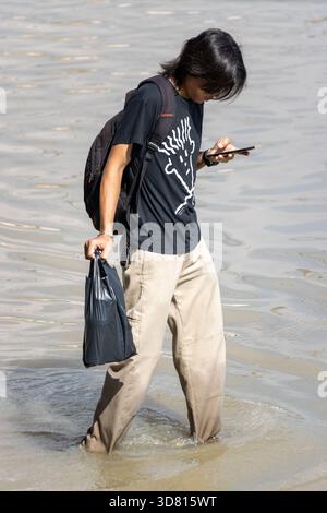 BANGKOK, THAÏLANDE, nov 09 2025, jeune homme patate dans l'eau dans une rue inondée et vérifie son téléphone Banque D'Images