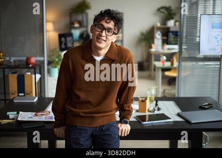 Portrait de jeune homme asiatique adulte portant des lunettes souriant et appuyé contre le bureau avec tablette numérique, documents et écran d'ordinateur dans l'espace de travail moderne Banque D'Images