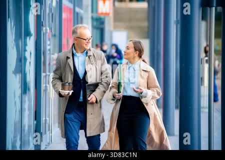 Homme d'affaires réussi et femme marchant dans la rue de la ville, souriant et parlant avec ordinateur portable et café. Concept moderne de travail d'équipe urbain et de succès. Banque D'Images