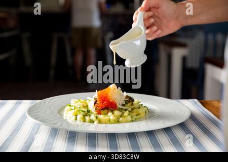 Le chef verse une sauce crémeuse sur un élégant hors-d'œuvre de fruits de mer gastronomique avec du caviar rouge et des légumes hachés sur une assiette blanche. Banque D'Images
