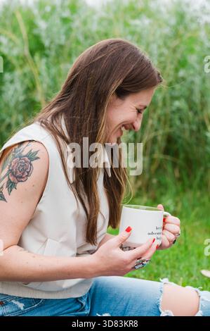 Femme riante tenant une tasse de café tout en étant assis sur l'herbe Banque D'Images