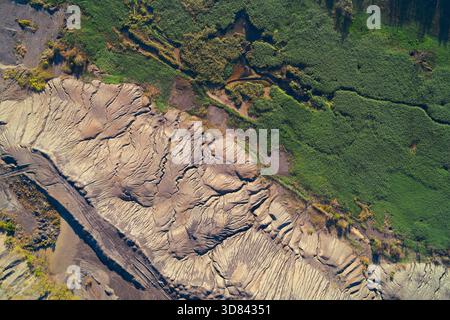 Vue aérienne abstraite d'une limite environnementale marquée. Les déchets industriels fortement érodés (résidus) contrastent fortement avec les roseaux verts luxuriants des zones humides, illustrant un conflit écologique grave. Banque D'Images