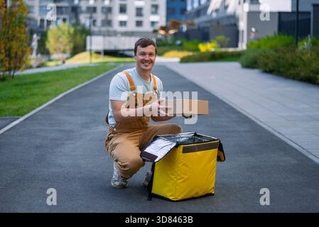 Livreur souriant et préparant un colis pour la livraison, le plaçant dans un sac isolant thermique jaune tout en travaillant à l'extérieur dans un environnement urbain moderne Banque D'Images