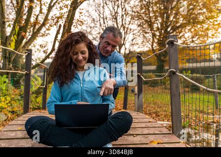 Couple mûr discutant de contenu sur un ordinateur portable tout en étant assis sur un pont en bois en automne Banque D'Images