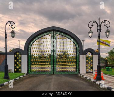 Entrée latérale de la mosquée Jame ASR Hassanil Bolkiah, Bandar Seri Begawan, Brunei Darussalam. Porte ornée de vert et d'or avec lampadaires et drapeau Brunei sous le ciel nuageux. Banque D'Images