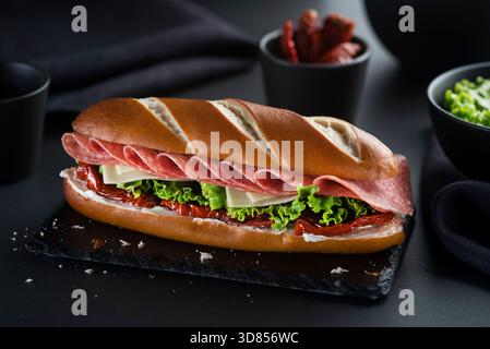 Le sandwich est rempli de couches de salami finement tranchées, de fromage, de tomate séchée et de laitue sur fond noir Banque D'Images
